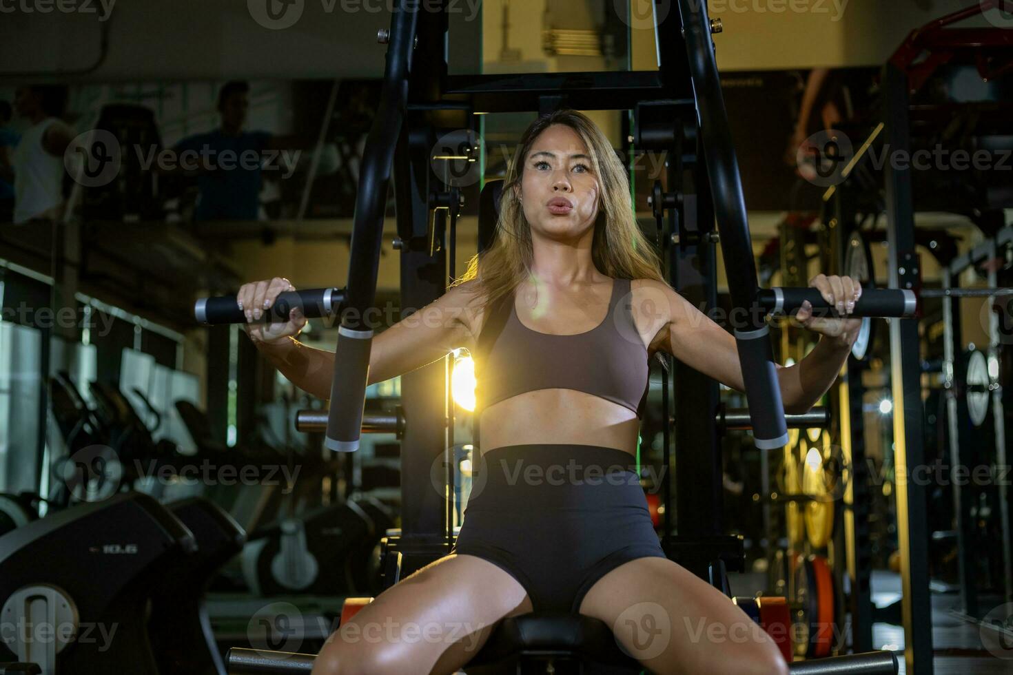 asiático mujer con fuerte y muscular cuerpo formación en cofre prensa máquina en el gimnasio para músculo edificio y peso levantamiento práctica con espalda ligero en oscuro tono foto