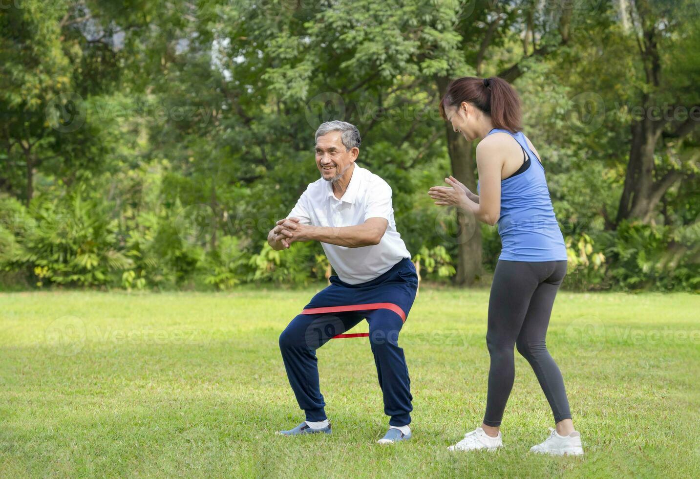 young woman trainer teaching a senior man doing exercise with elastic band,smiling woman applauding beside him,legs stretch exercise for rehabilitation muscles in older adult,concept of elderly health photo