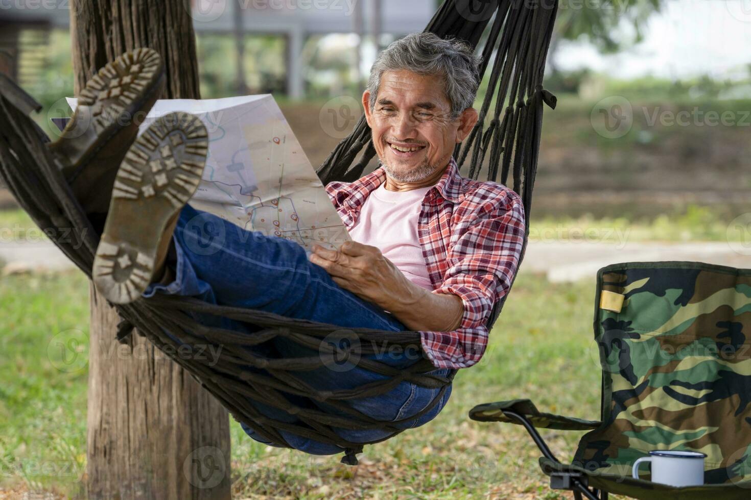 happy senior male with grey hair and beard wears checkered shirt relax at resort garden on summertime,mature man laying on rope hammock,smile and look at a paper map,concept of travel,camping,resting photo