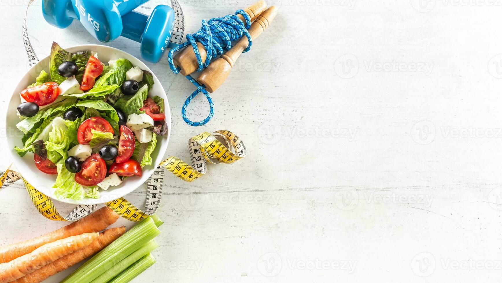Healthy fresh salad with tomatoes surrounded with exercise equipment, carrtot celery and measuring tape - top of view photo