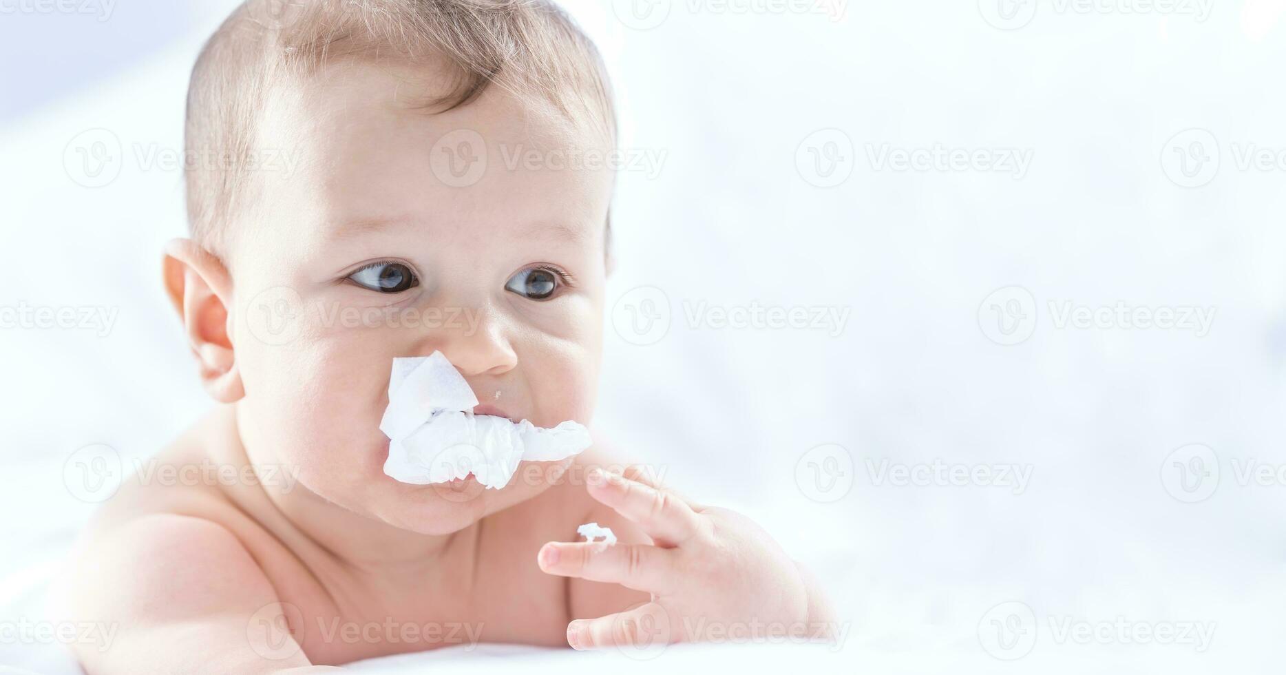 Funny portrait of cute toddler boy lying on bed and trying to eat a paper napkin. Newborn child in bedroom. Baby lies on his tummy and raises the head photo