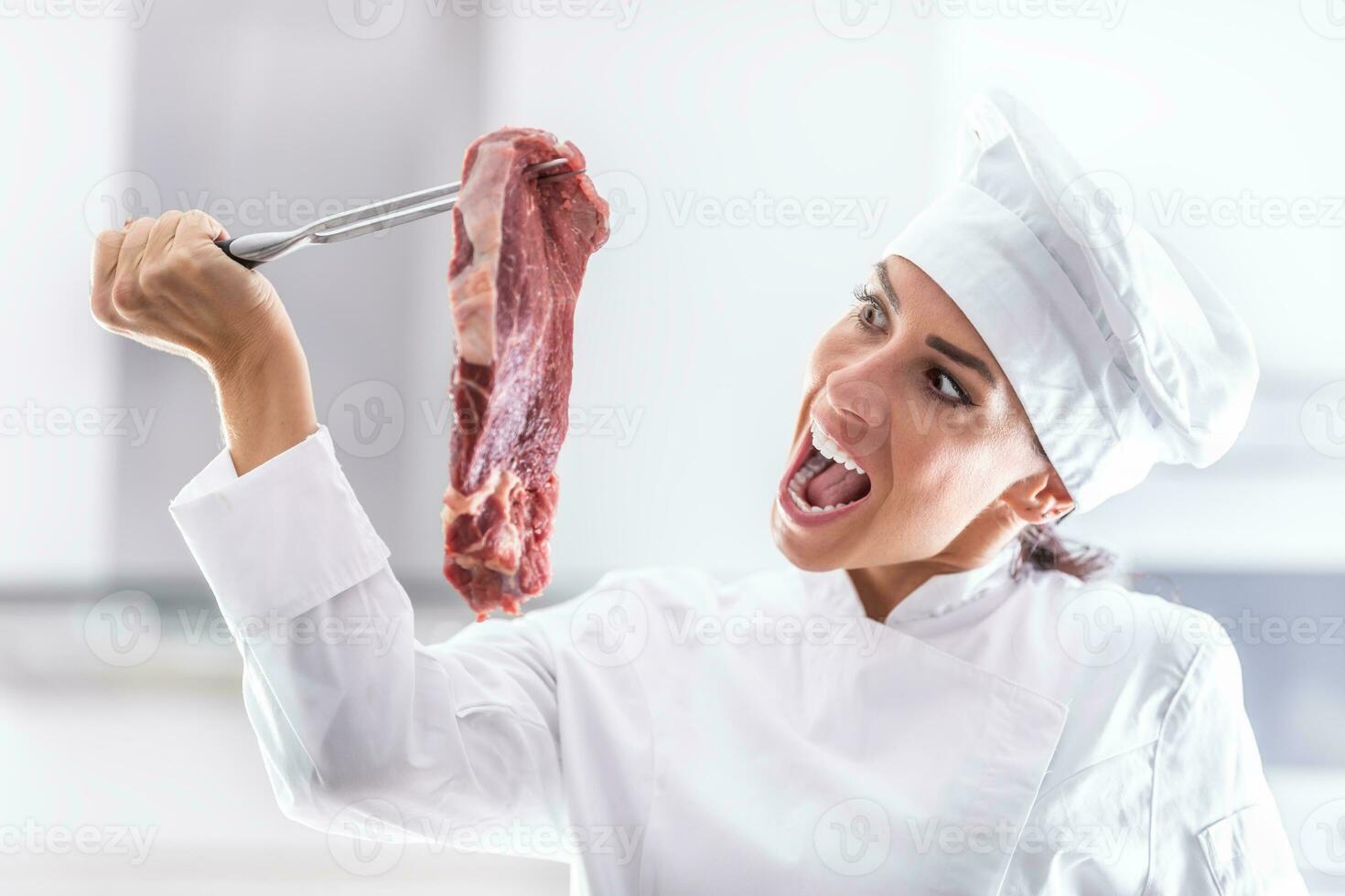 Female chef in rondon pretends wanting to bite into a raw cut of red meat she holds on a fork photo