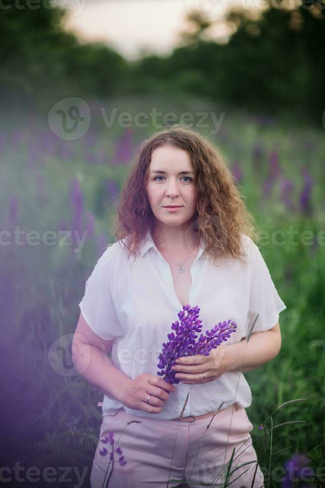 Young woman stands in white shirt in field of purple and pink lupins. Beautiful young woman with curly hair outdoors on a meadow, lupins blossom. Sunset or sunrise, bright evening light photo