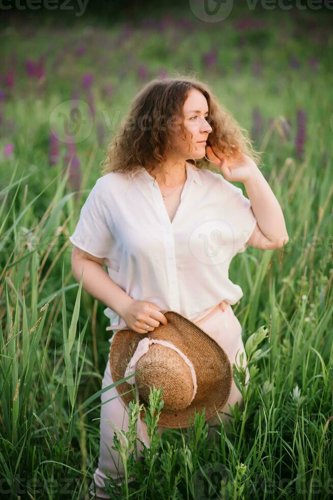 Young woman stands in white shirt in field of purple and pink lupins. Beautiful young woman with curly hair and hat outdoors on a meadow, lupins blossom. Sunset or sunrise, bright evening light photo