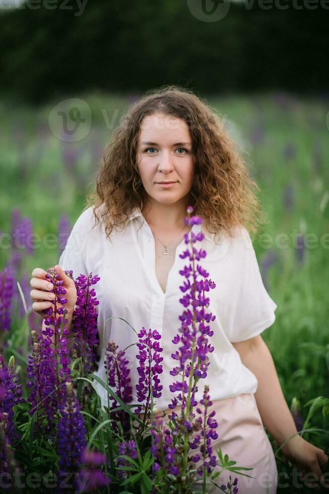 joven mujer soportes en blanco camisa en campo de púrpura y rosado altramuces hermosa joven mujer con Rizado pelo al aire libre en un prado, altramuces florecer. puesta de sol o amanecer, brillante noche ligero foto