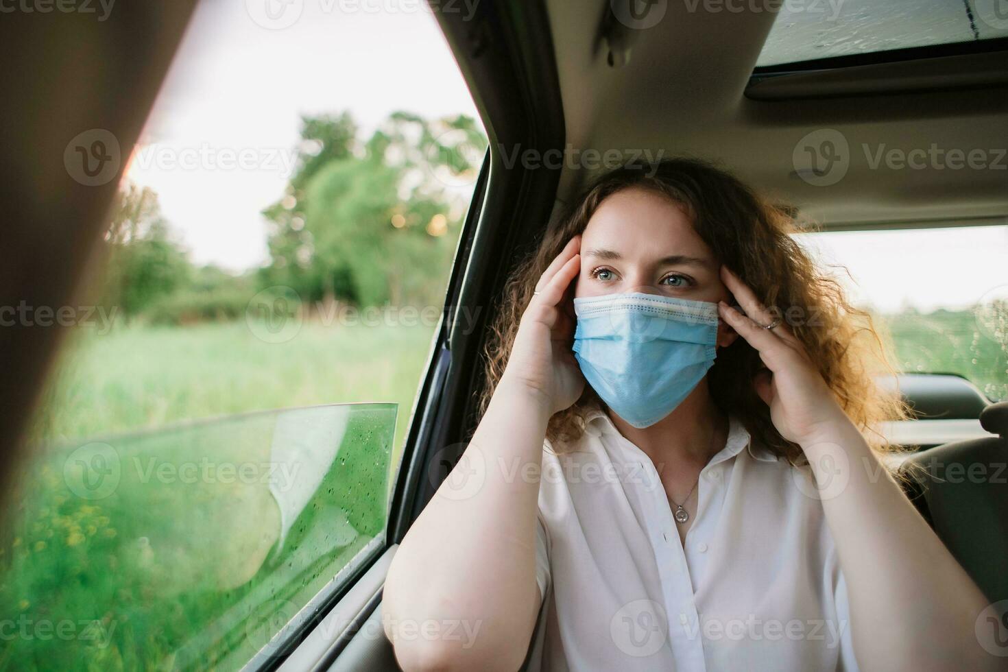 elegante joven carly mujer en médico máscara tener un dolor de cabeza mientras sentado en espalda asiento de coche en natural antecedentes con puesta de sol foto