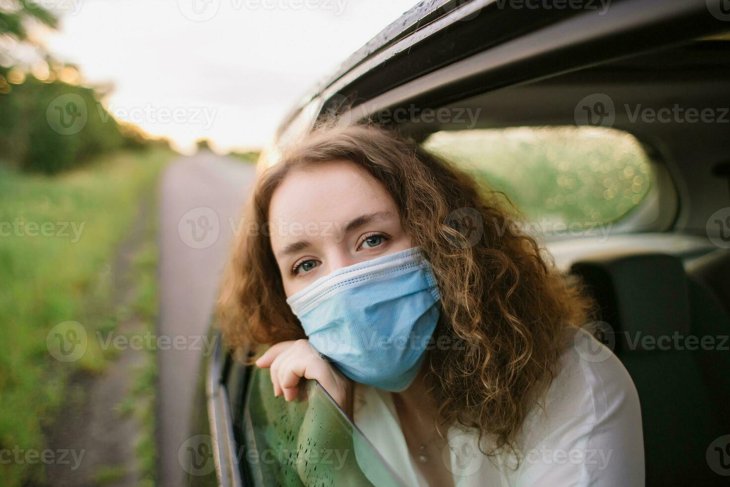 elegante joven carly mujer en médico máscara mirando fuera ventana mientras sentado en espalda asiento de coche en borroso antecedentes con puesta de sol foto