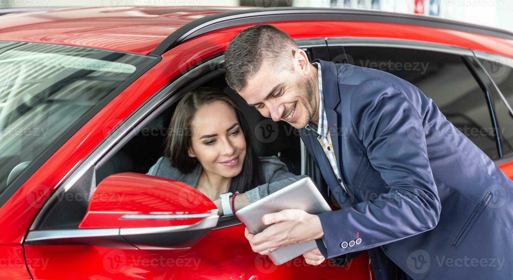 Car dealership employee with tablet shows woman intending to buy a red car she sits in technical features photo