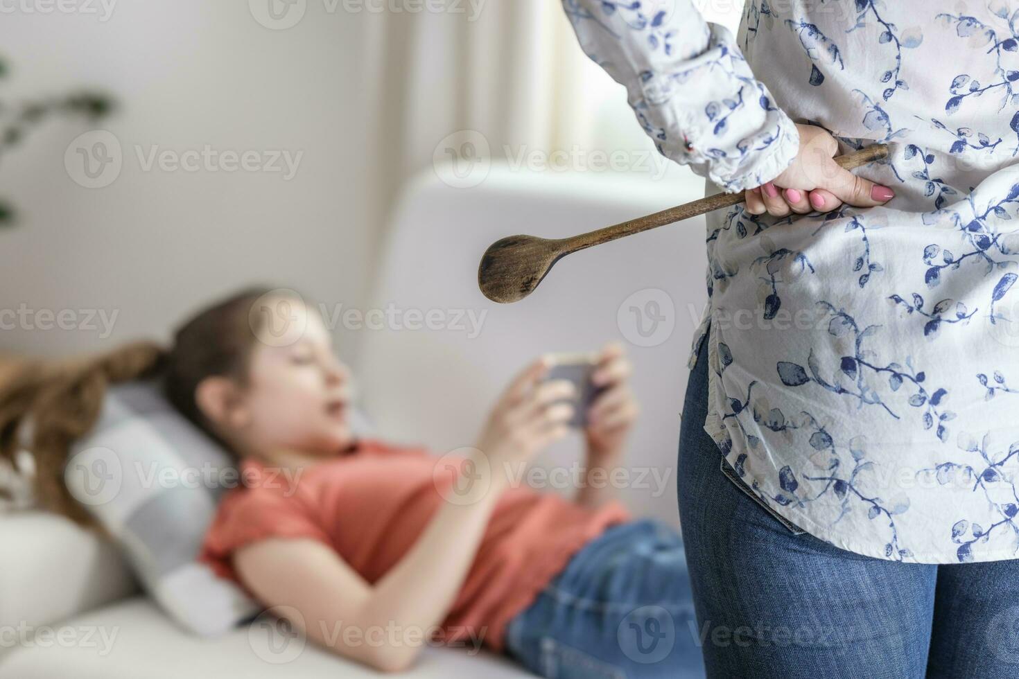 Adolescent girl ignores her mother standing upset with a wooden spoon next to her and keeps being on her mobile phone photo