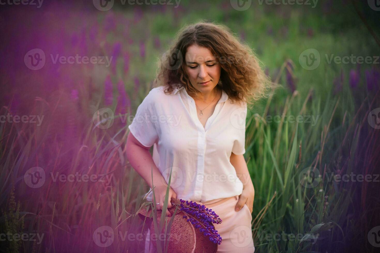 Young woman stands in white shirt in field of purple and pink lupins. Beautiful young woman with curly hair and hat outdoors on a meadow, lupins blossom. Sunset or sunrise, bright evening light photo