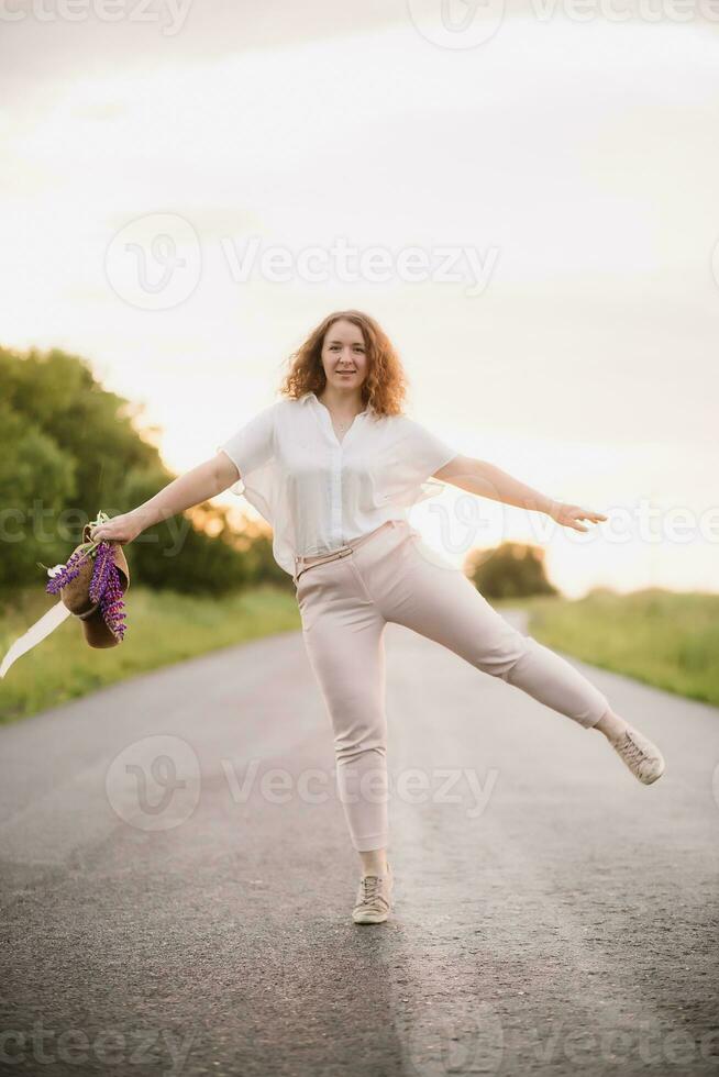 joven mujer soportes en blanco camisa en la carretera con púrpura y rosado altramuces hermosa joven mujer con Rizado pelo con ramo de flores de altramuces puesta de sol o amanecer, brillante noche ligero foto