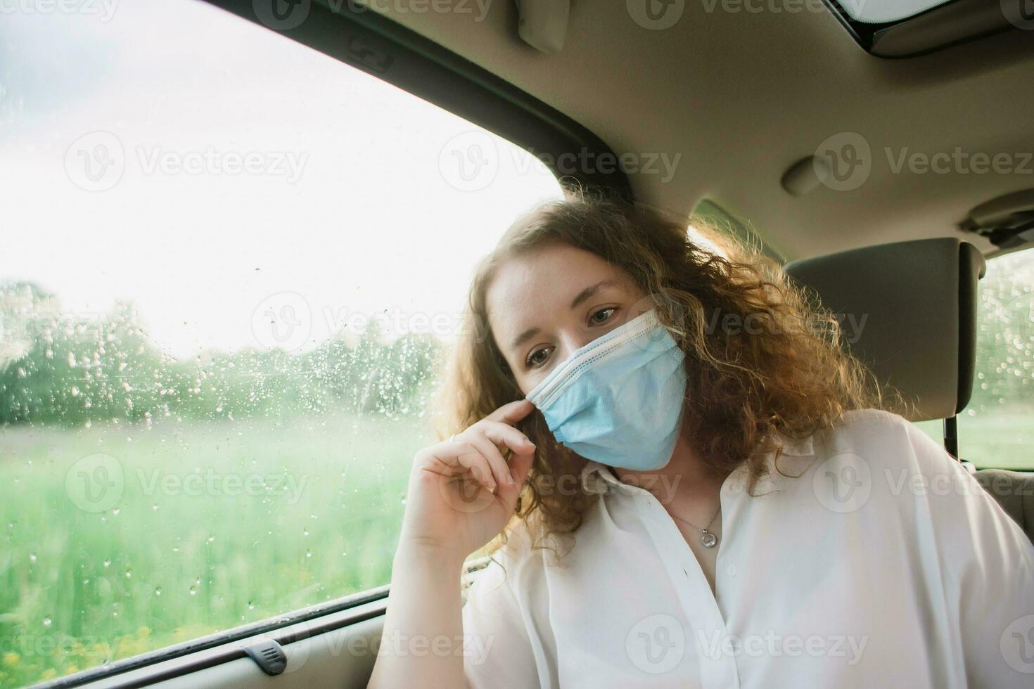 elegante joven carly mujer en médico máscara sentado en espalda asiento de coche en borroso antecedentes con puesta de sol. foto