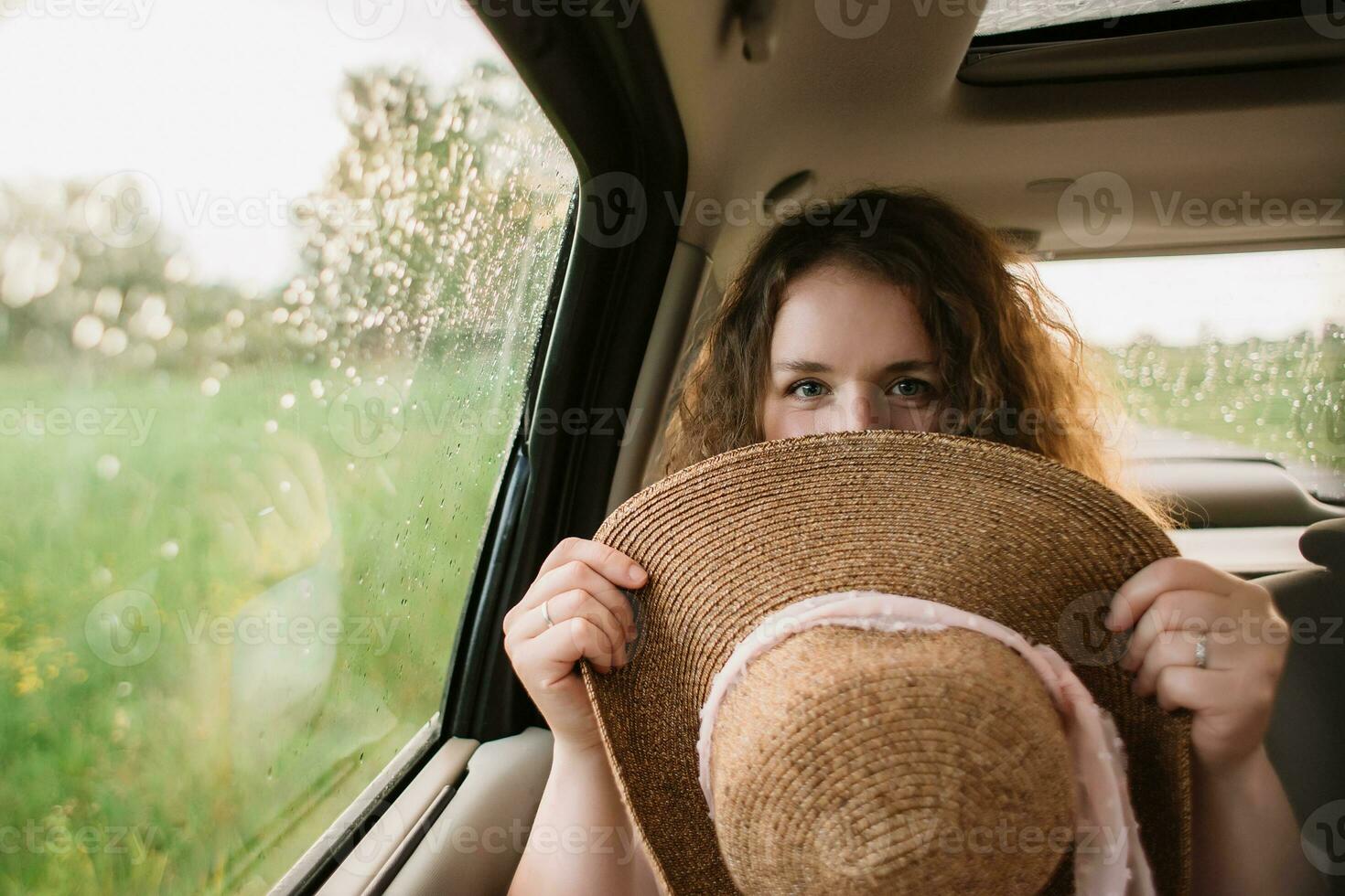 Cheerful positive curly young woman in casual wear sitting in automobile backseat with fastened seatbelt with hat. photo