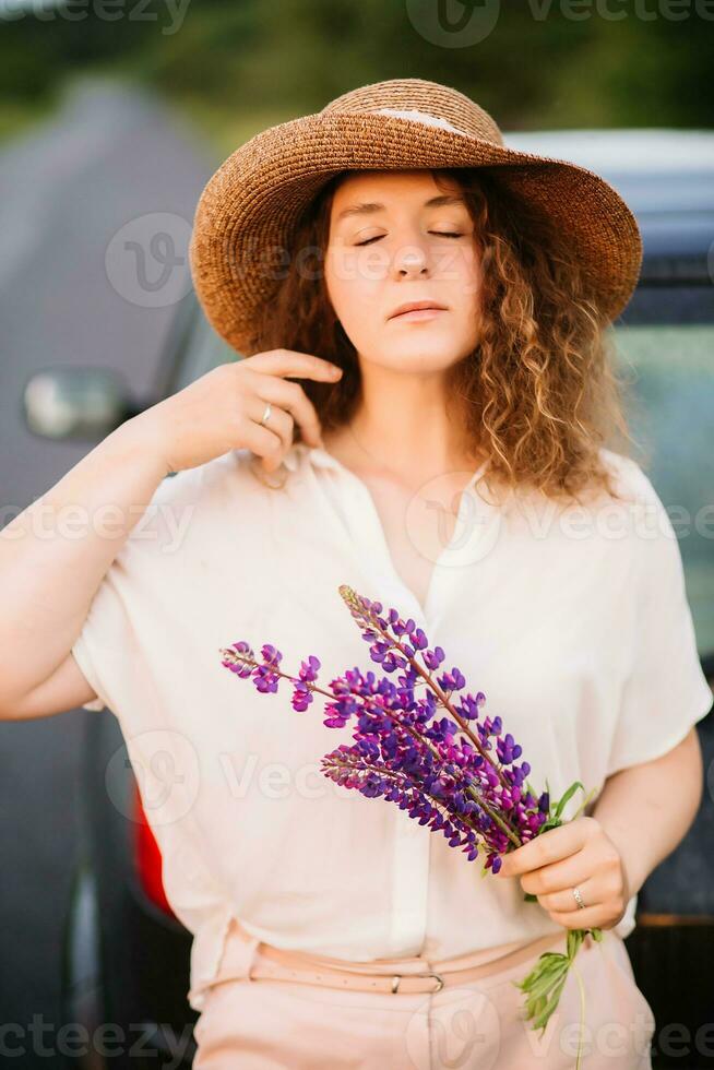 joven mujer soportes en blanco camisa cerca coche con púrpura y rosado altramuces hermosa joven mujer en sombrero con Rizado pelo con ramo de flores de altramuces puesta de sol o amanecer, brillante noche ligero foto