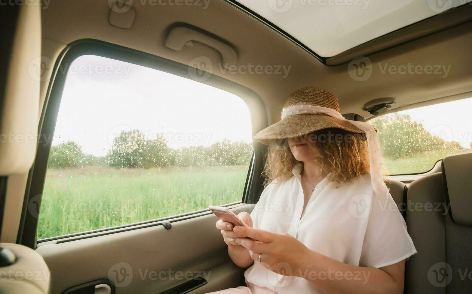 Cheerful positive curly young woman in casual wear with hat sitting in automobile backseat with fastened seatbelt and using phone. photo