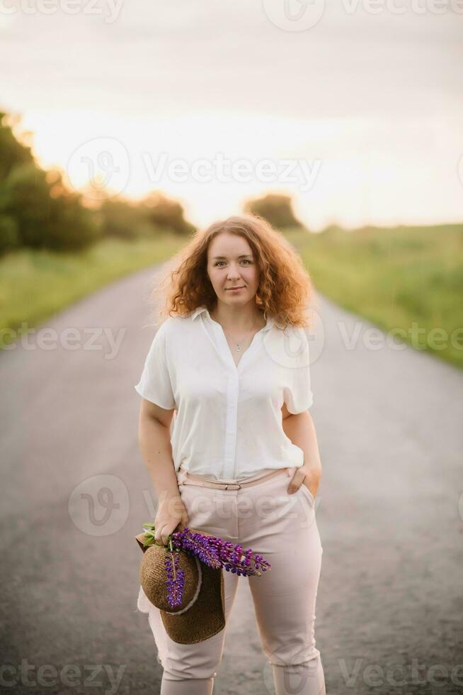 Young woman stands in white shirt on road with purple and pink lupins. Beautiful young woman with curly hair with bouquet of lupins. Sunset or sunrise, bright evening light photo