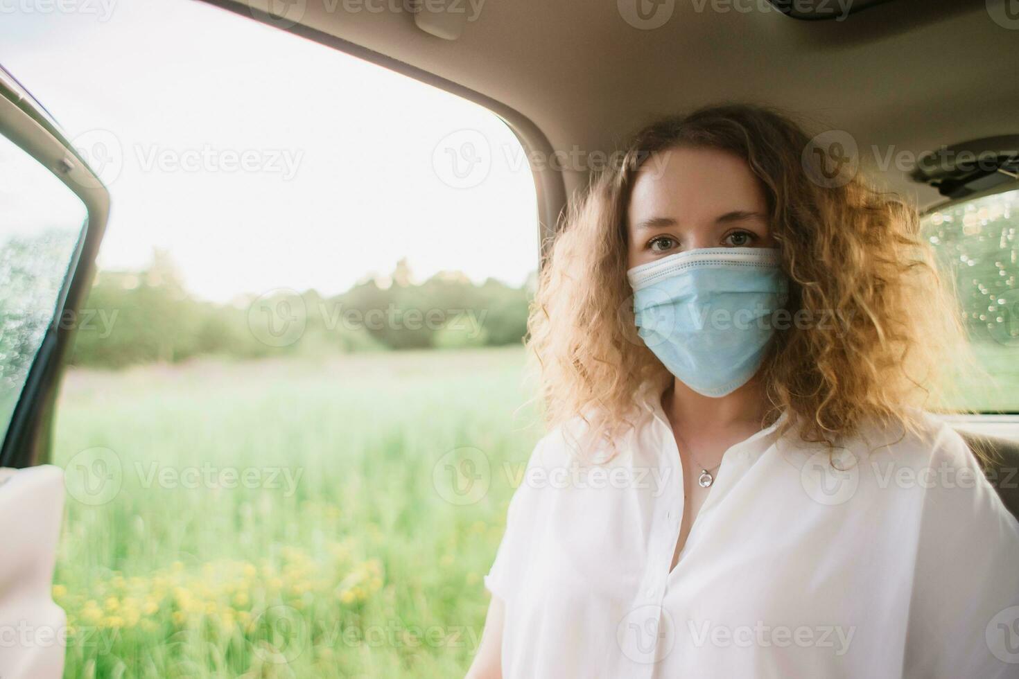 elegante joven carly mujer en médico máscara sentado en espalda asiento de coche en borroso antecedentes con puesta de sol foto