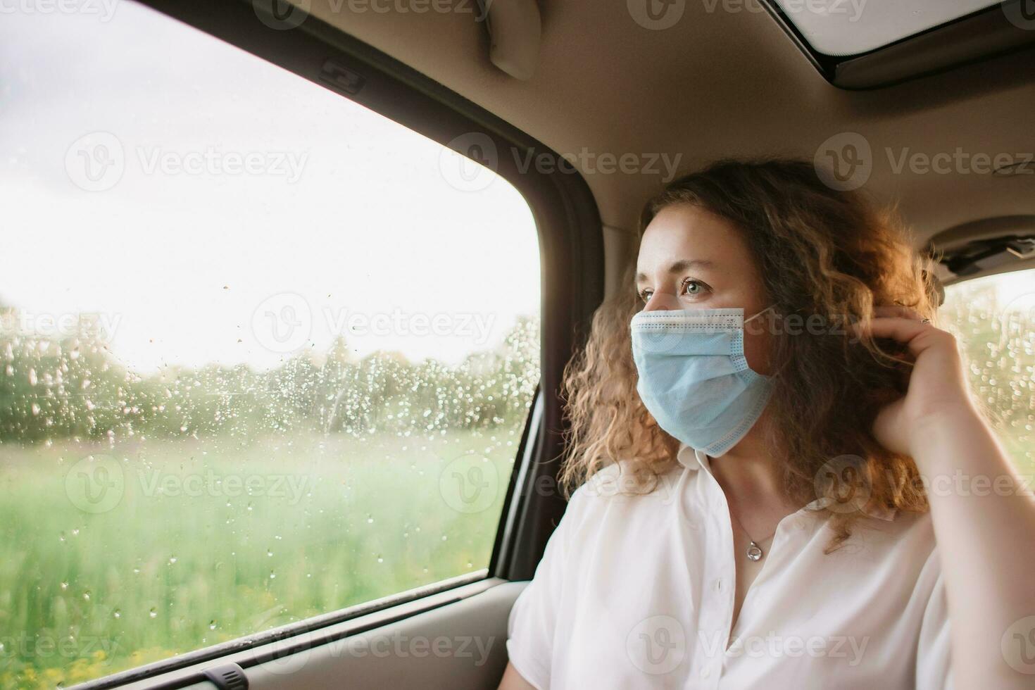 elegante joven carly mujer en médico máscara mirando fuera ventana mientras sentado en espalda asiento de coche en borroso antecedentes con puesta de sol foto