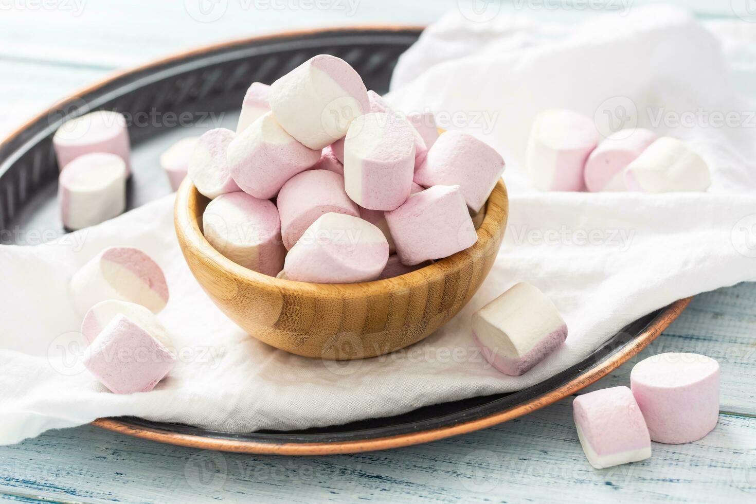 Wooden bowl full of pink and white marshmallows with some scattered around on a white table cloth, dark tray and white wooden table photo