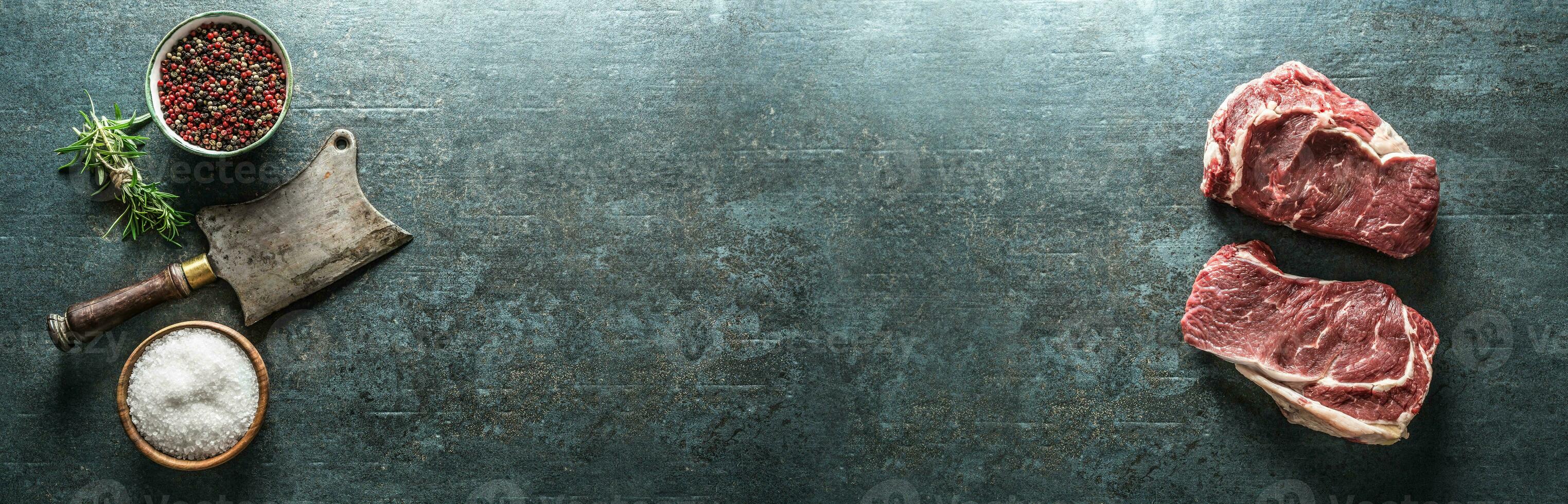 Two aged raw beef steaks with meat cleaver and salt pepper on dark concrete background - Top of view panoramic banner photo