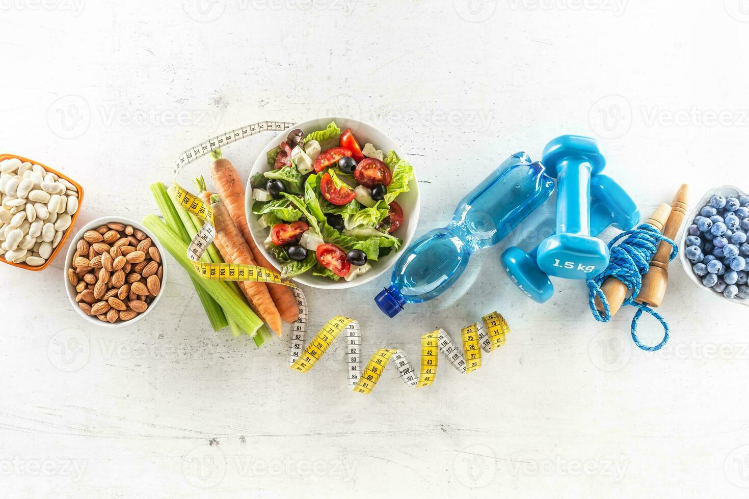 Healthy fresh salad with tomatoes surrounded with exercise equipment, carrtot celery and measuring tape - top of view photo