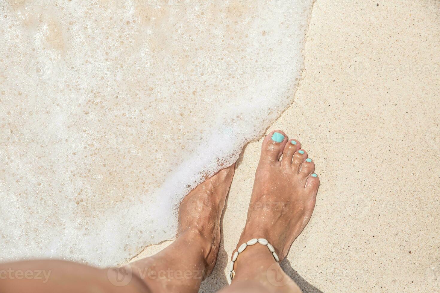 Bare feet of a woman with turquoise nail polish standing on a sandy beach washed out by foamy wave of water from the sea photo