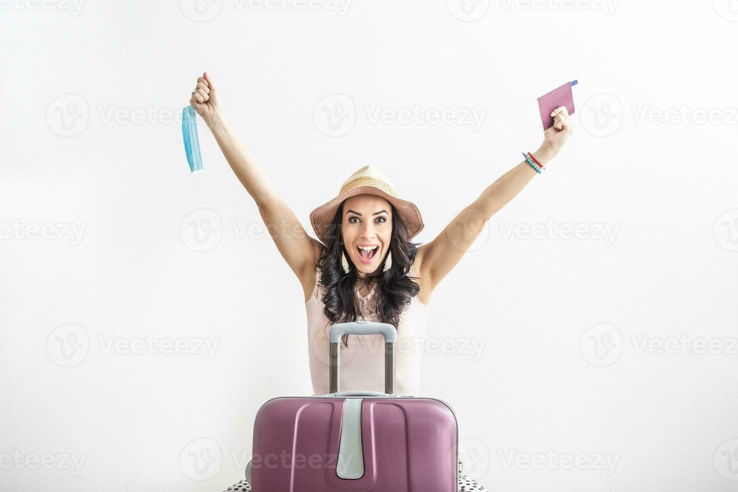 Happy female traveler stands in front of her suitcase, arms up as restrictions ease, holding passport in one and face mask in the other hand photo