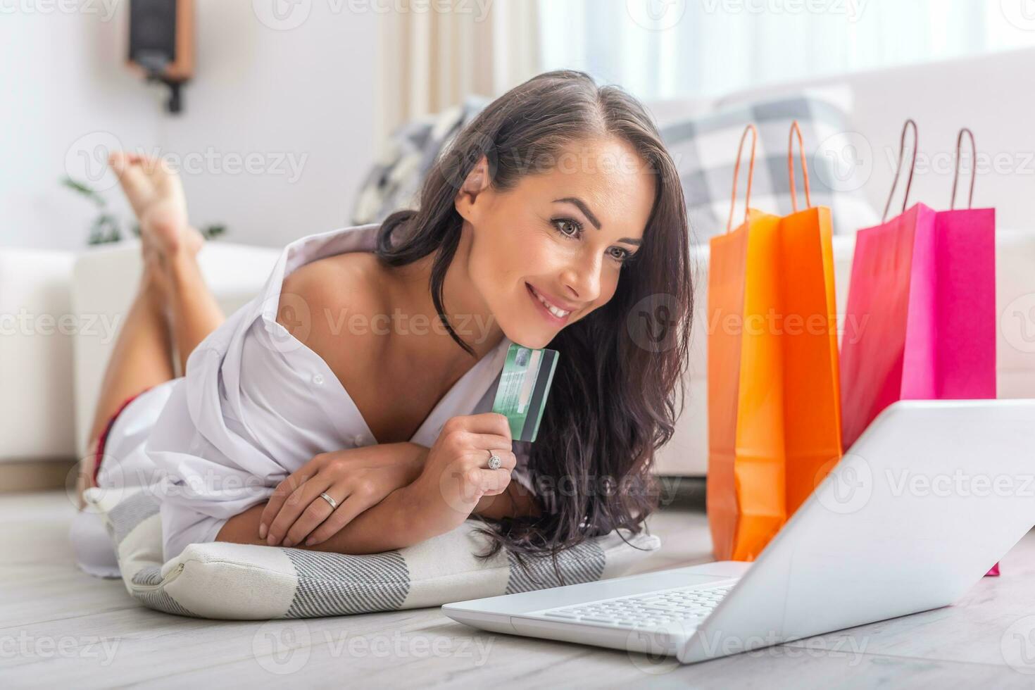 Sexy brunette female shopping online while lying on the floor in light living room, with a pillow under her elbows, a debit card in her hand, smiling. Orange and pink paper shopping bags next to her photo