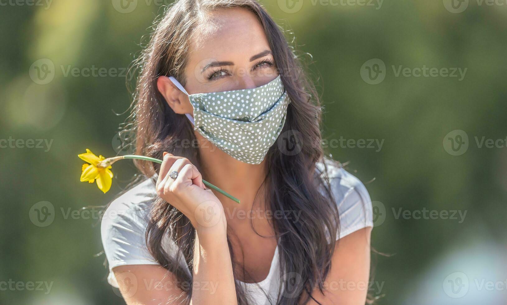Brunette girl in the nature wears facemask smiling, holding a daffodil in her hand photo
