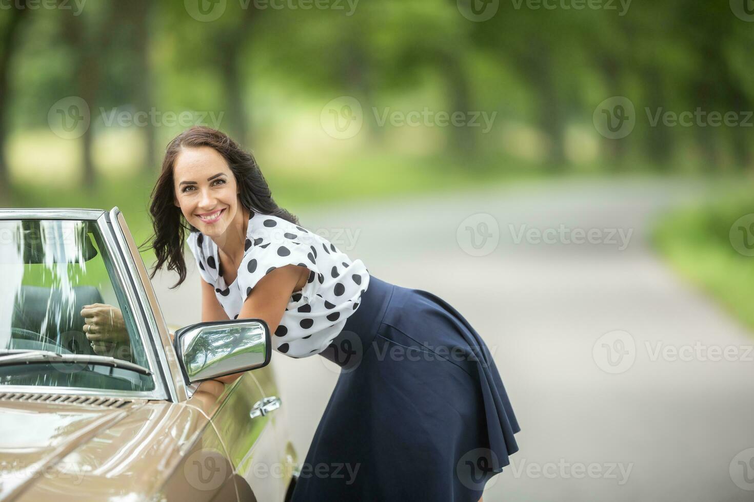 hermosa niña en falda y blusa sonrisas a el cámara, propensión hacia adelante a un convertible coche foto