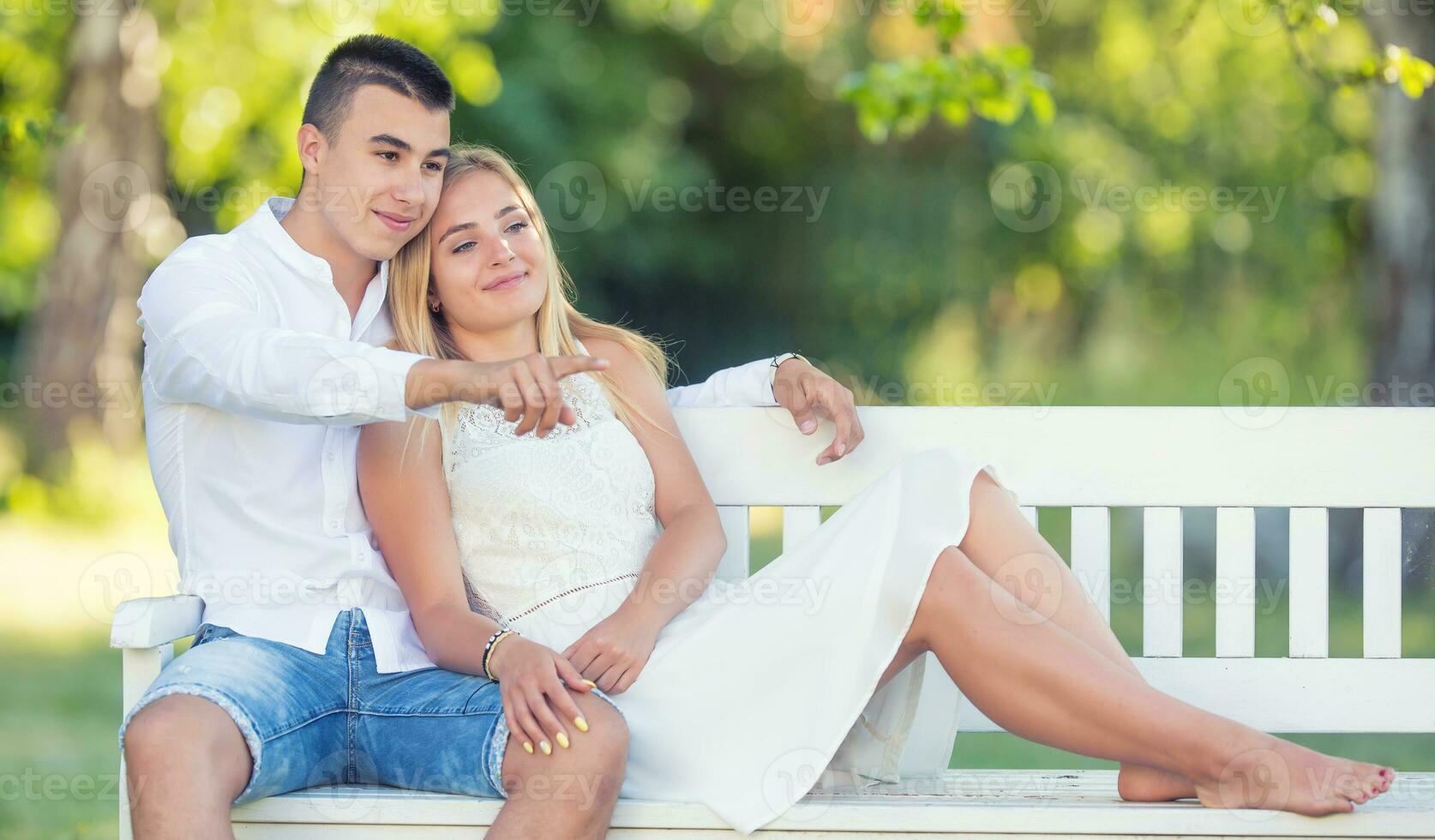Young couple dressed in light clothes sitting on a white bench in a park looking the same direction. Boy pointing with a finger, while the couple is smiling photo
