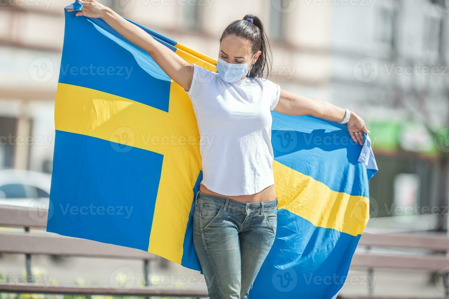 Brunetter standing outdoors looks at the Swedish flag she holds, with a face mask on photo