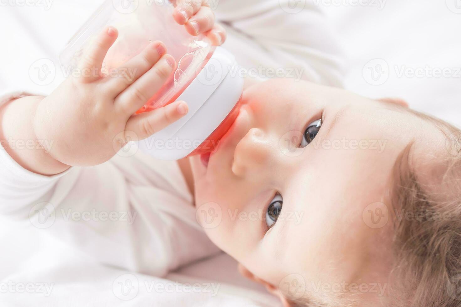 bebé chico acostado en cama y Bebiendo té desde su el plastico botella. foto