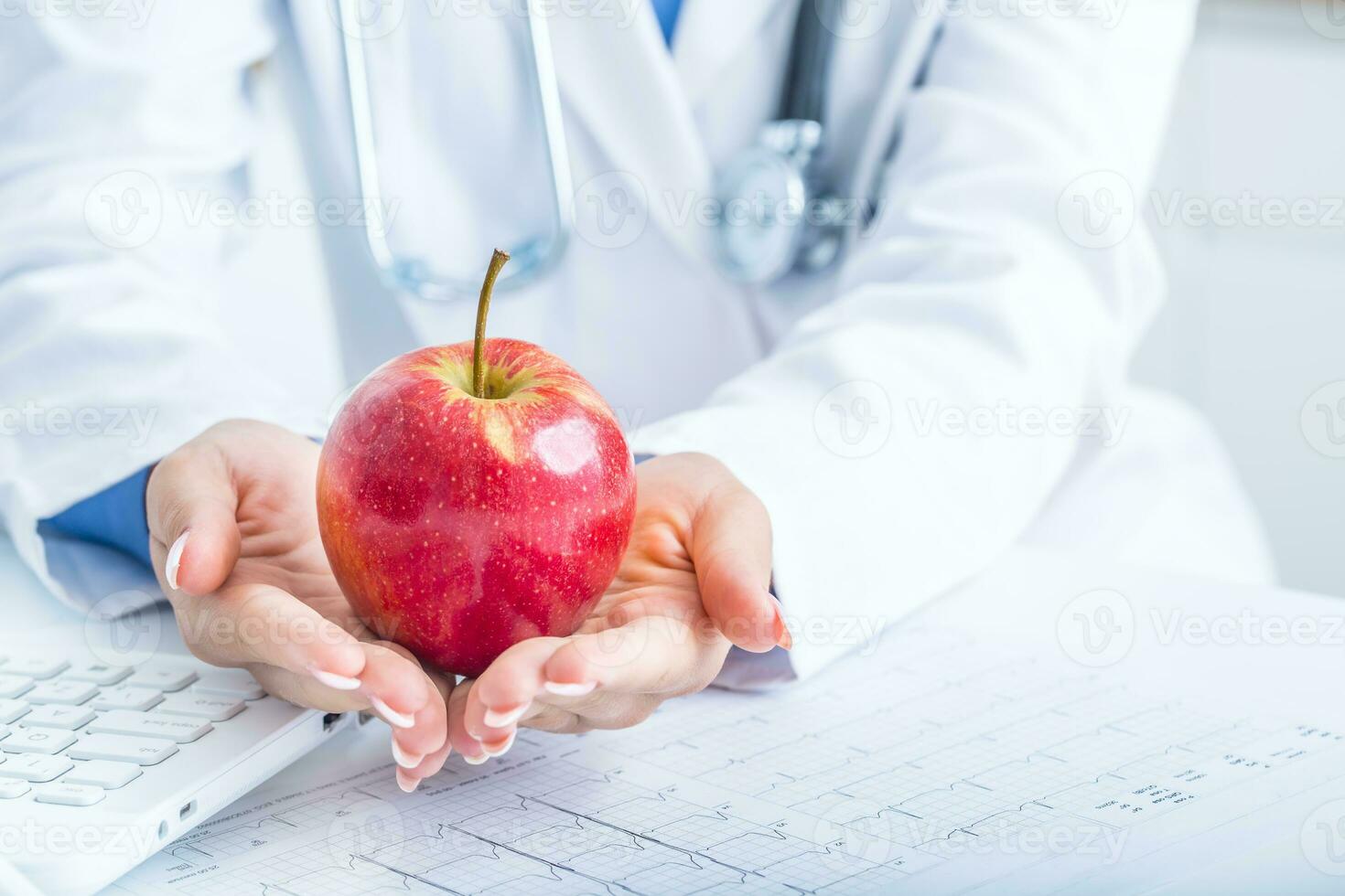 Woman doctor in ambulance holds red apple in hands. On desk laptop and EKG diagram photo
