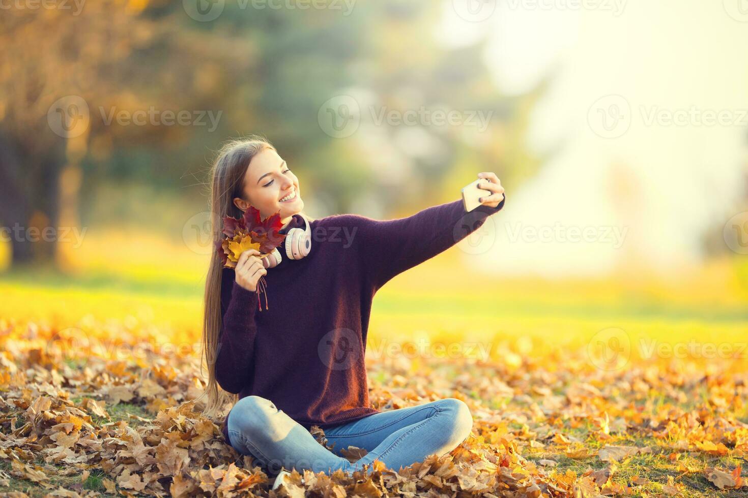 Portrait of happy young girl with headphones and smartphone in autumn park listenig music or makes selfie photo