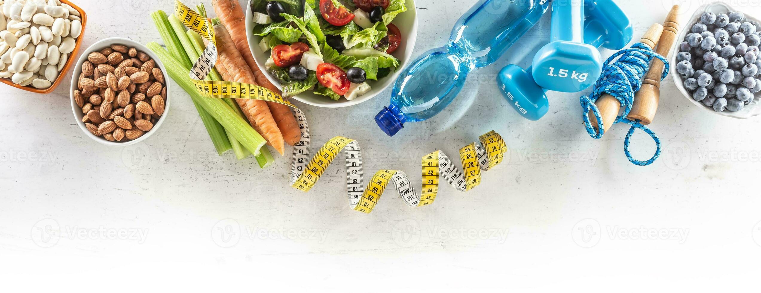 Healthy fresh salad with tomatoes surrounded with exercise equipment, carrtot celery and measuring tape - top of view photo