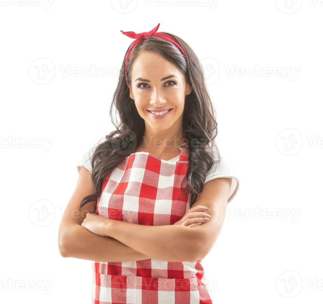 bueno mirando sonriente morena cocinar en pie con confianza en aislado blanco antecedentes con brazos cruzado en su pecho. vestido en rojo y blanco comprobado delantal y un rojo cinta en su pelo foto