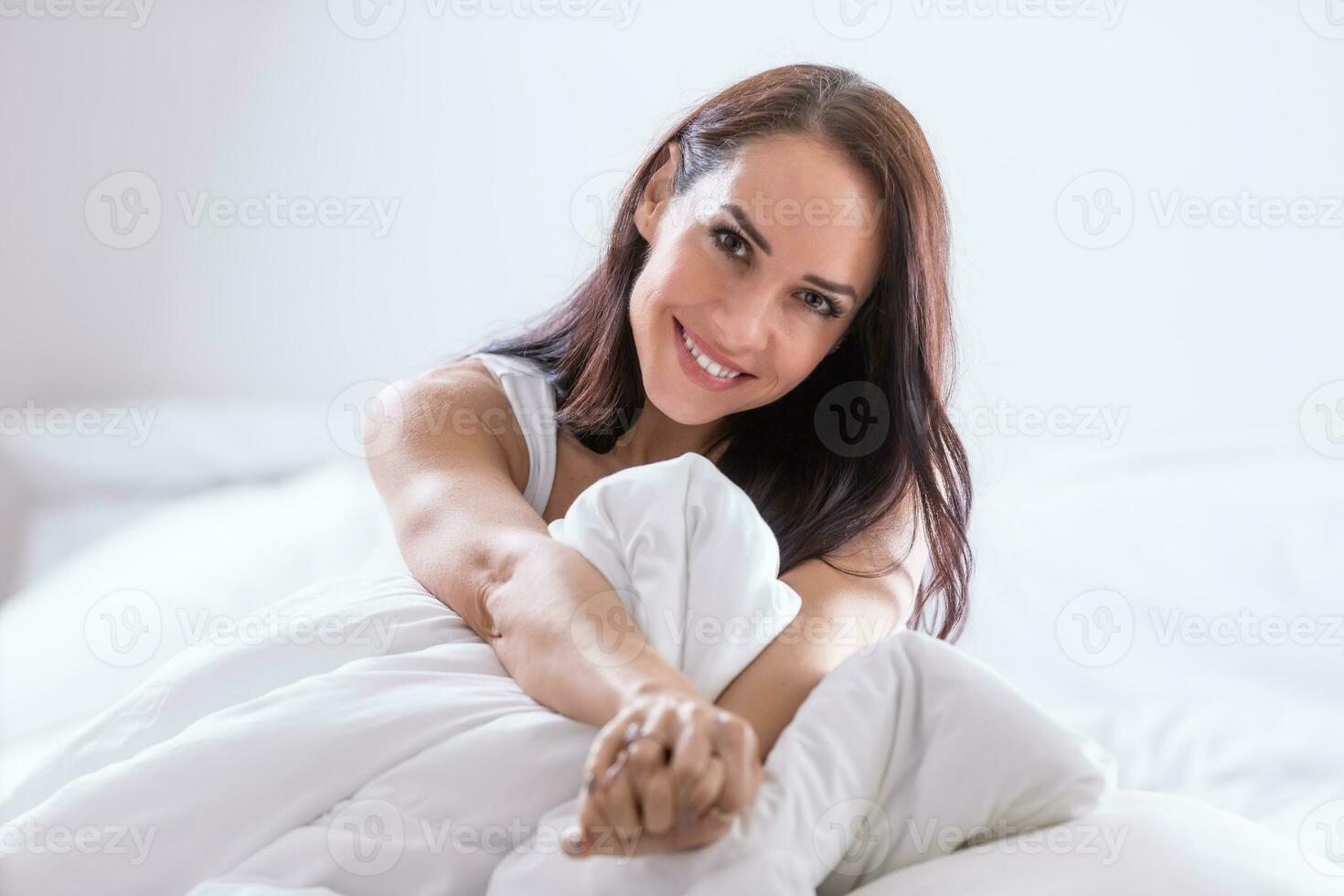 guapo mujer se sienta cubierto en blanco plumón estirar su brazos, sonriente foto