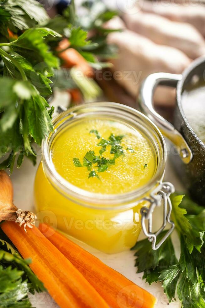Chicken broth in a glass jar raw chicken legs carrot onion celery herbs garlic and fresh vegetables photo