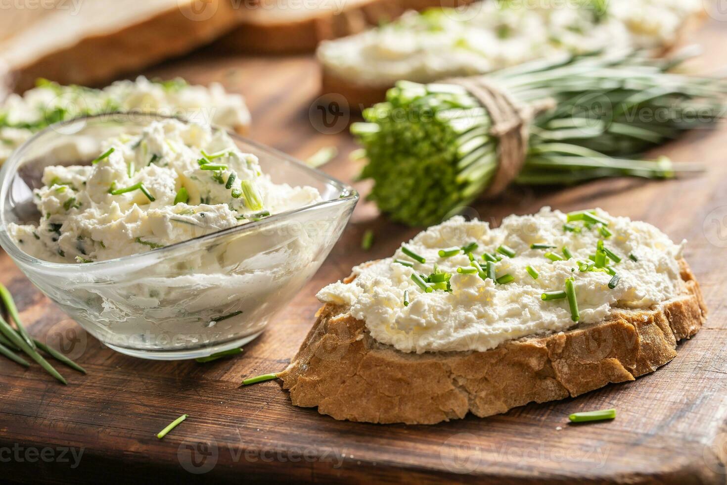 A bowl of homemade cream cheese spread with chopped chives surrounded by bread slices with spread and a bunch of freshly cut chives photo