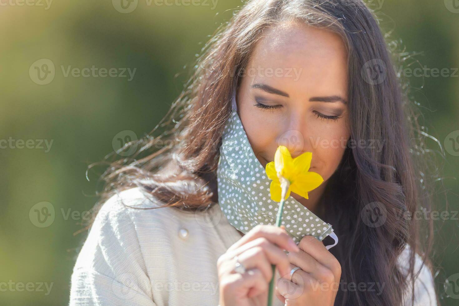 Pretty woman enjoys the smell of a flower with half of her face mask off, having closed eyes on a sunny day in the nature photo