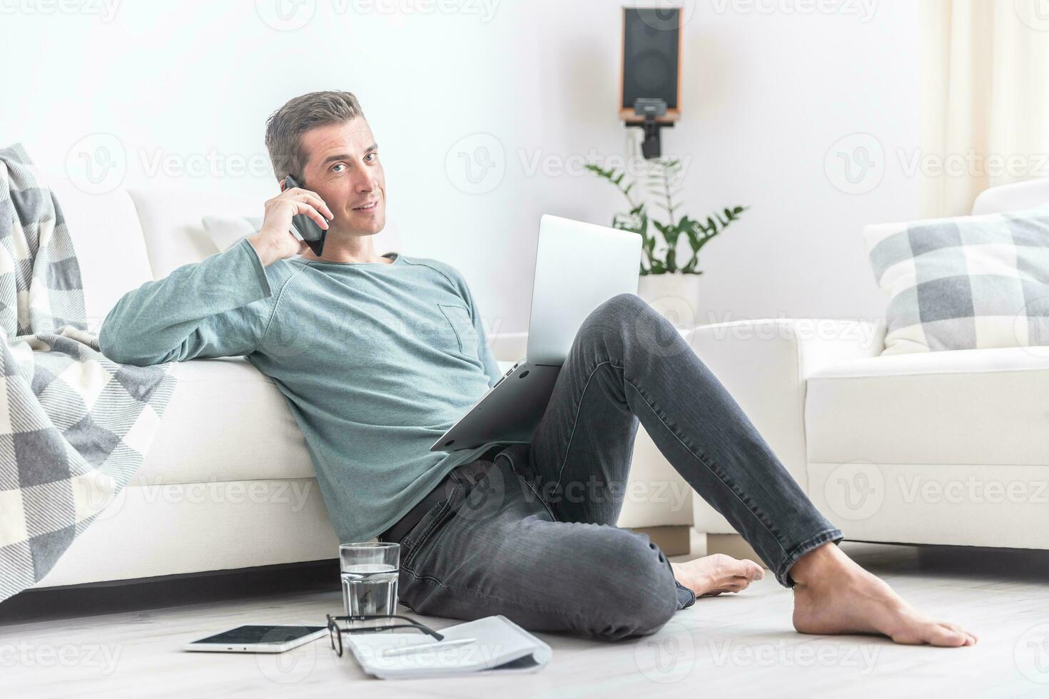 casual ropa vistiendo hombre se sienta en el piso en el departamento, vocación en un célula teléfono con un cuaderno en frente de él foto