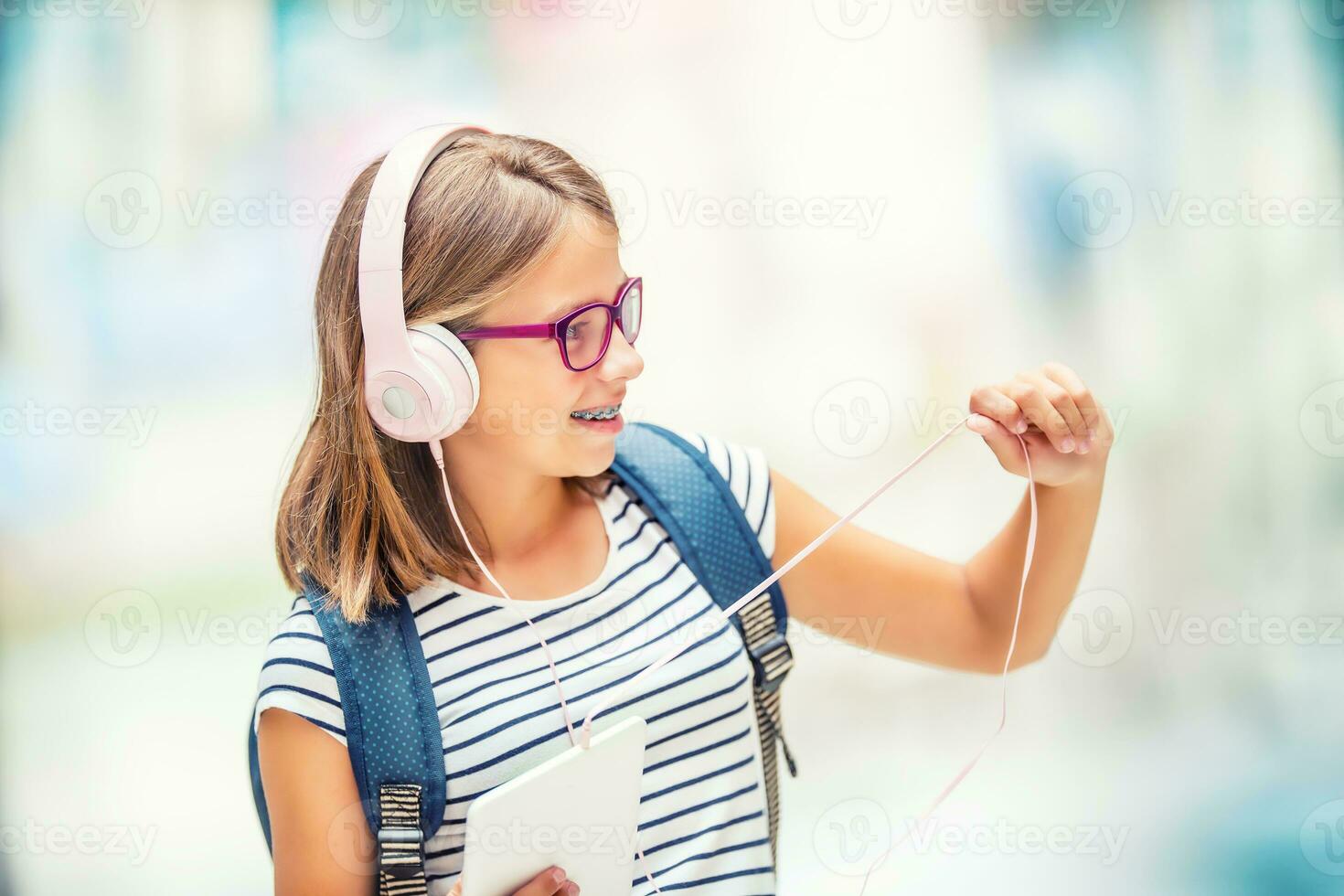 Schoolgirl with bag, backpack. Portrait of modern happy teen school girl with bag backpack headphones and tablet. Girl with dental braces and glasses. photo