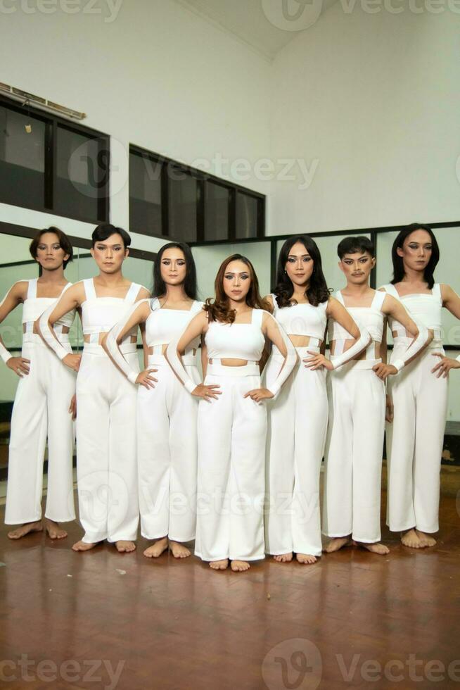 a group of Asian dancers stand side by side in white costumes in a room with a dance floor photo