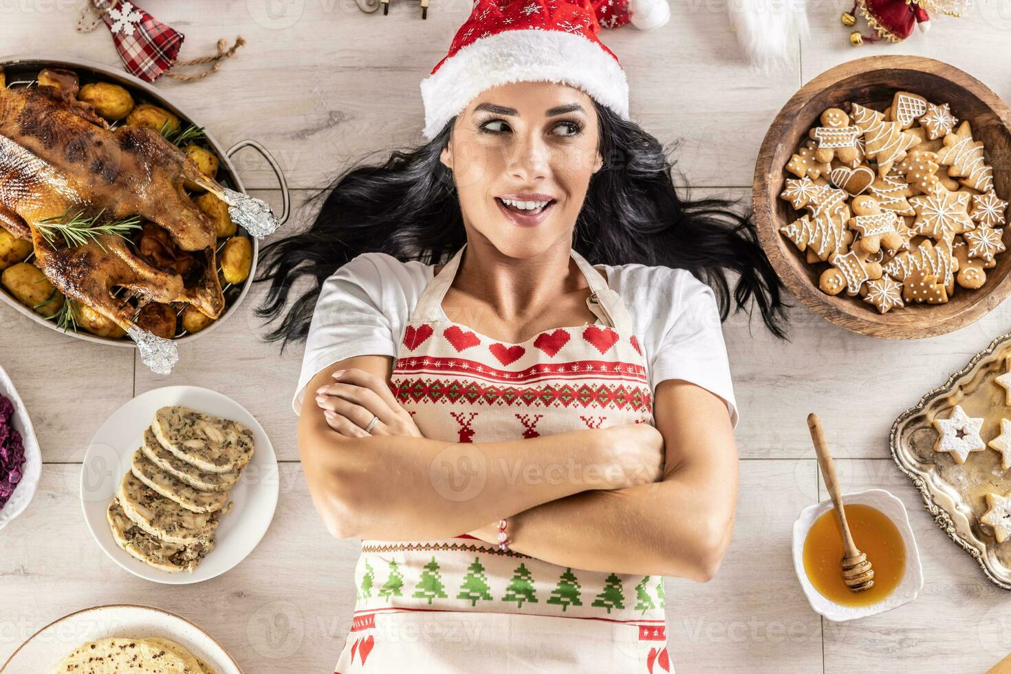 Satisfied female cook in a Christmas apron and Santa hat,lying on the floor, surrounded by gingerbread, Linz cakes, on the other side, roast goose or turkey with side dishes photo