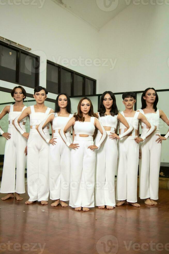 a group of Asian dancers stand side by side in white costumes in a room with a dance floor photo