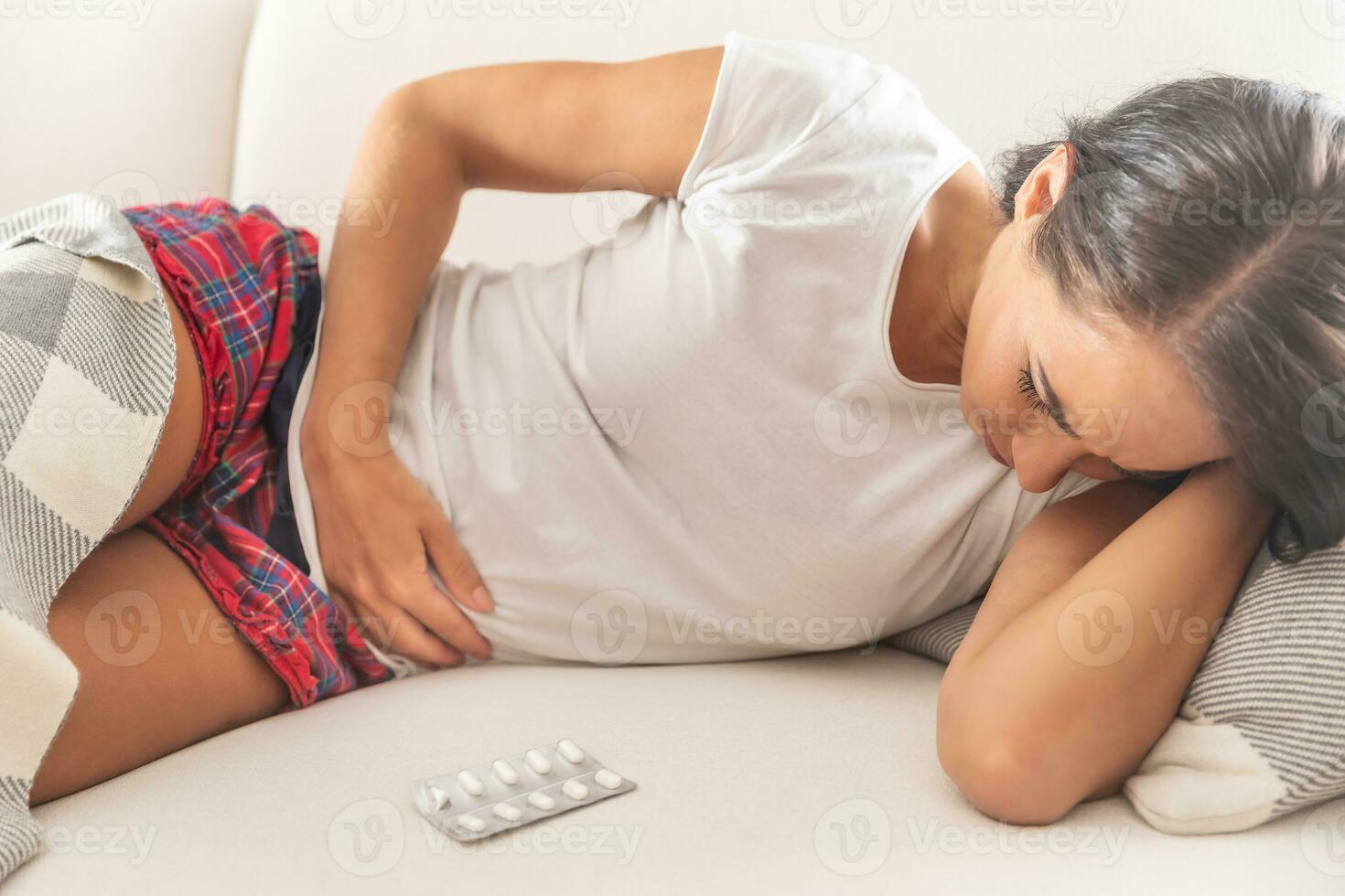 Girl with pain in lower abdomen lying on her side on a couch with hand below her head and other hand holding her belly. Partially covered by a blanket with a blister pack of pills in front of her photo