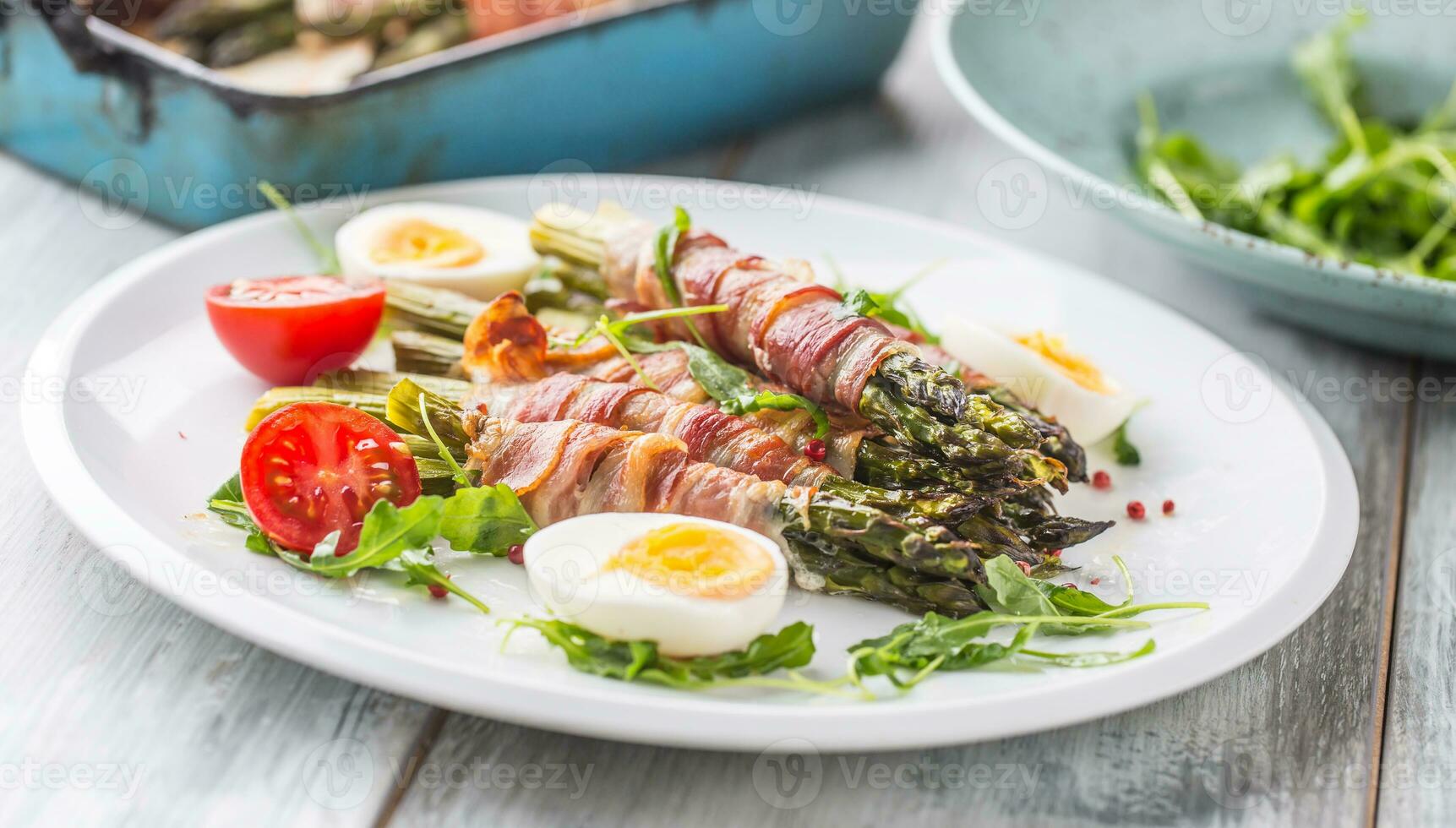 Fresh asparagus wrapped in bacon  on a white plate with arugula tomatoes and eggs photo