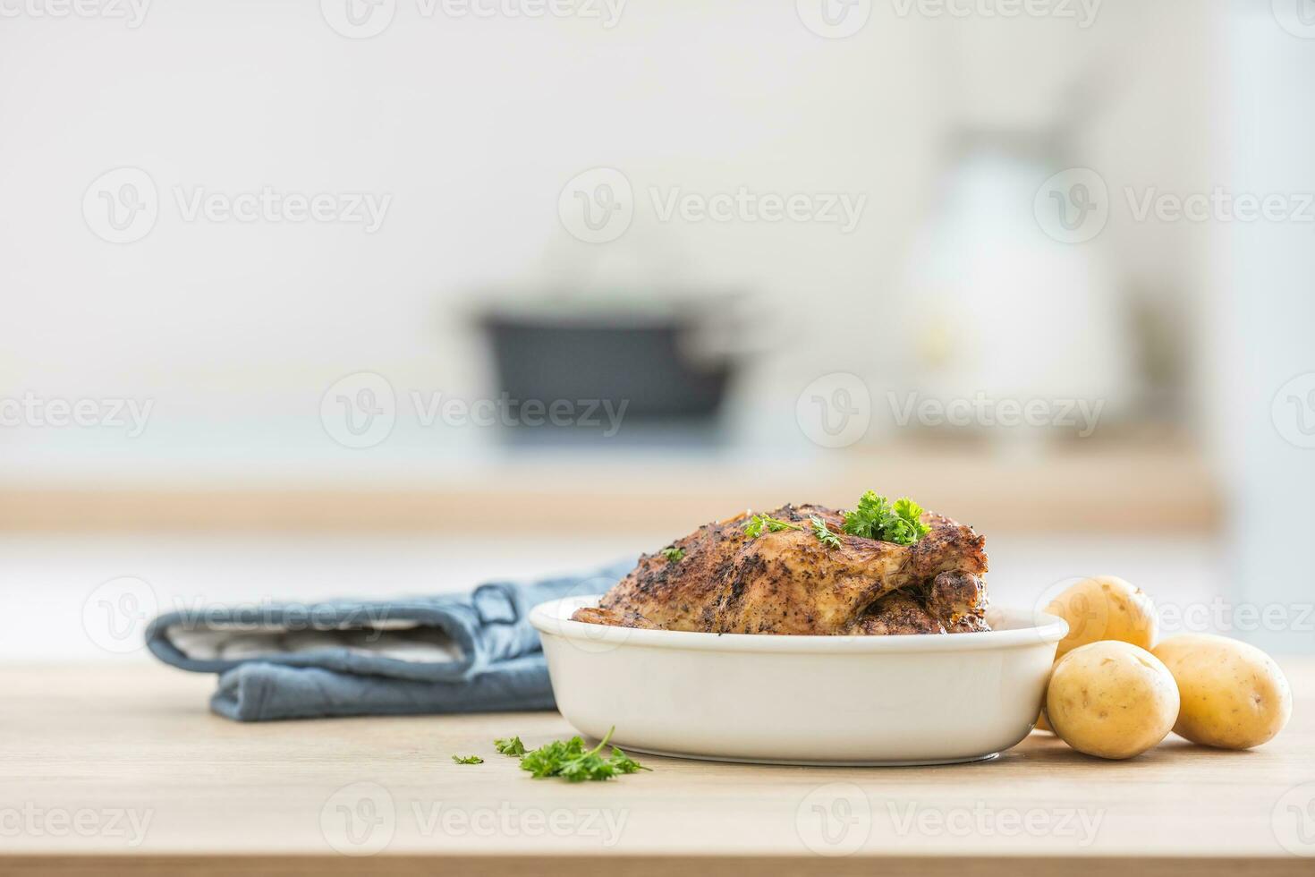 Whole chicken roasted in white dish with raw potatoes on kitched table. Home interior in the background photo