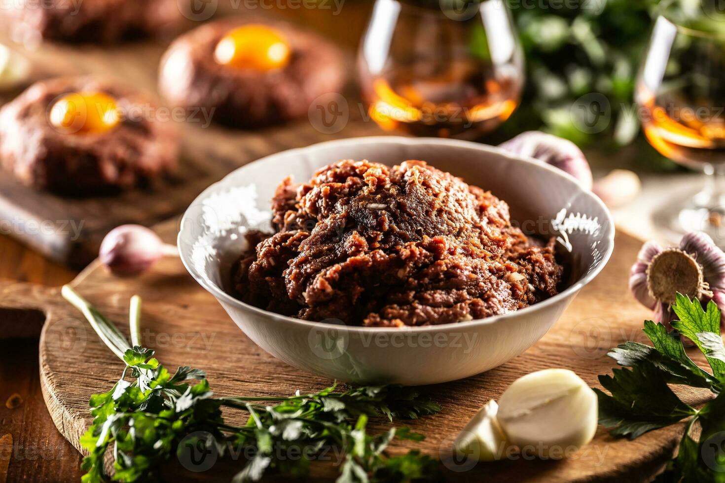 White bowl filled with raw meat ready to prepare delicious steak tartare with savory egg yolks. Already prepared tartares in the backgroud with rum and herbs as a decoration photo